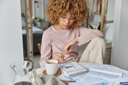 Serious curly haired woman manages household family budget calculates expenditures takes care of finances and savings sits at table with receipts dressed in domestic clothes poses at home office