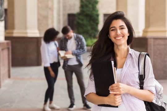 Happy beautiful student girl with backpack holding folders at university university and friends background. Student loans concept